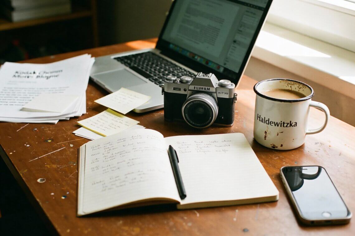 Arbeitsplatz eines Rentner-Bloggers mit Fujifilm X-T30 Kamera, aufgeschlagenem Notizbuch mit handschriftlichen Notizen, Laptop, Emaille-Kaffeetasse mit Haldewitzka-Logo, Smartphone und beschriebenen Papieren auf Holztisch – typischer Alltag im Ruhestand beim Bloggen und Fotografieren