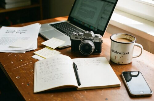 Arbeitsplatz eines Rentner-Bloggers mit Fujifilm X-T30 Kamera, aufgeschlagenem Notizbuch mit handschriftlichen Notizen, Laptop, Emaille-Kaffeetasse mit Haldewitzka-Logo, Smartphone und beschriebenen Papieren auf Holztisch – typischer Alltag im Ruhestand beim Bloggen und Fotografieren