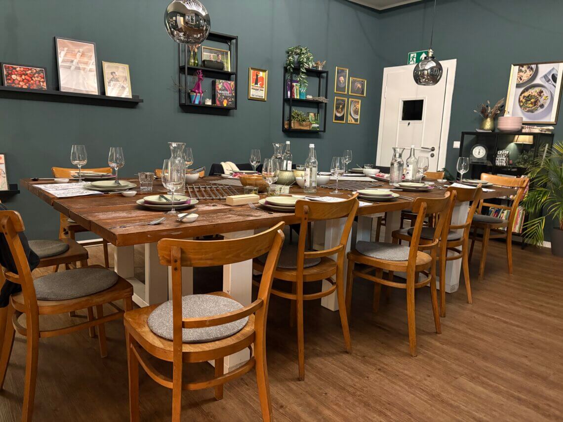 Alt-Text: Festlich gedeckter Tisch mit Holzstühlen in gemütlichem Kochstudio, petrolgrüne Wand, bereitet für gemeinsames Essen nach Kochkurs.