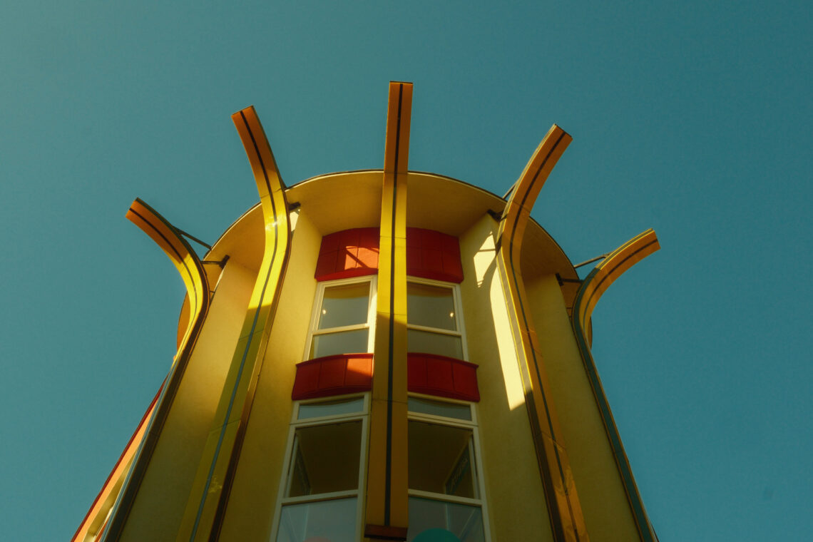 Gelber Rundturm mit roten Fensterelementen und geschwungenen Metallstreben, von unten fotografiert vor türkisblauem Himmel – architektonische Detailaufnahme im Gegenlicht in der Fußgängerzone von Wuppertal