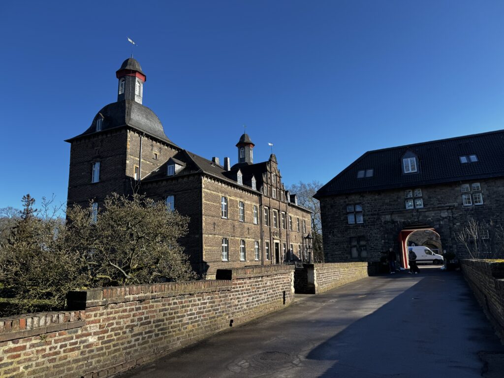 Schloss Hugenpoet in Essen-Kettwig mit markanter Backsteinfassade, Rundturm mit Wetterfahne und Torhaus bei klarem Winterhimmel