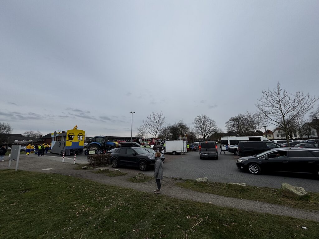 Blick auf den Parkplatz vor dem Archäologischen Park Xanten während eines Karnevalsumzugs: links ein gelber Karnevalswagen in Form einer Minion-Figur mit kostümierten Teilnehmern, rechts Fahrzeugverkehr und eine einzelne Frau als Beobachterin, kahle Bäume und bewölkter Winterhimmel