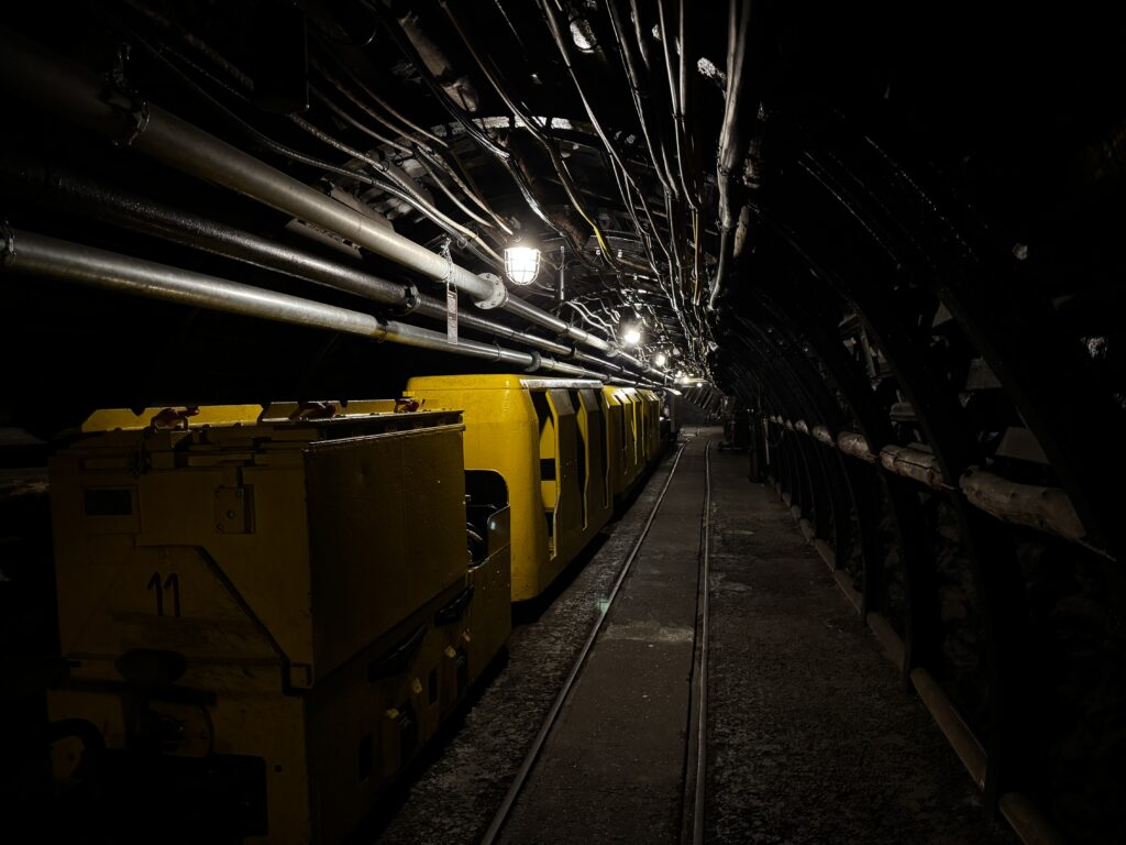 Blick in einen beleuchteten Stollen im Deutschen Bergbau-Museum Bochum mit Schienen auf dem Boden und technischer Ausstattung an den Wänden.