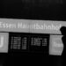 Silhouetten zweier Personen vor dem beleuchteten Abfahrtstafel-Schild am Essen Hauptbahnhof, Schwarz-Weiß-Streetfotografie