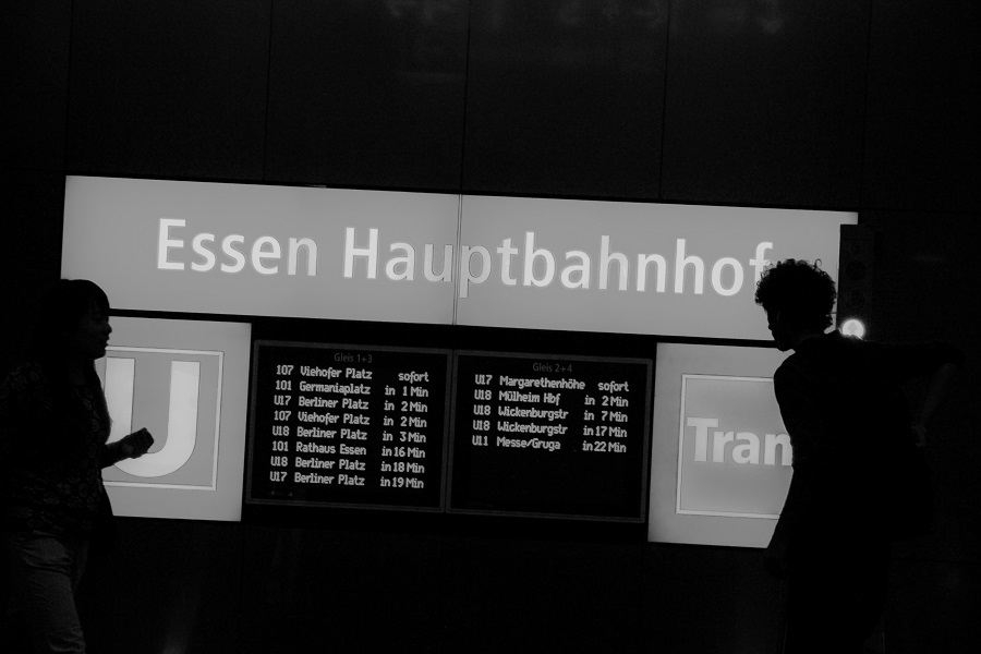 Silhouetten zweier Personen vor dem beleuchteten Abfahrtstafel-Schild am Essen Hauptbahnhof, Schwarz-Weiß-Streetfotografie