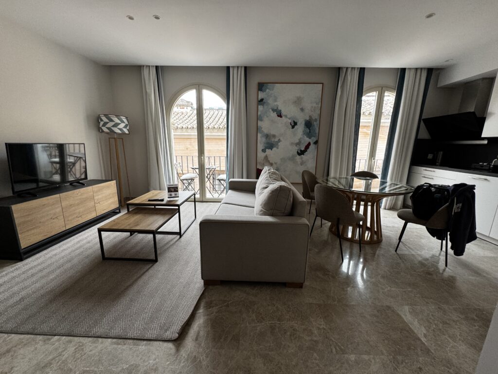 Modernes Hotelzimmer oder Ferienapartment mit hellem Wohnbereich, Bogenfenstern mit Blick auf spanische Dachziegel, abstraktem Wandbild und zeitgenössischer Einrichtung