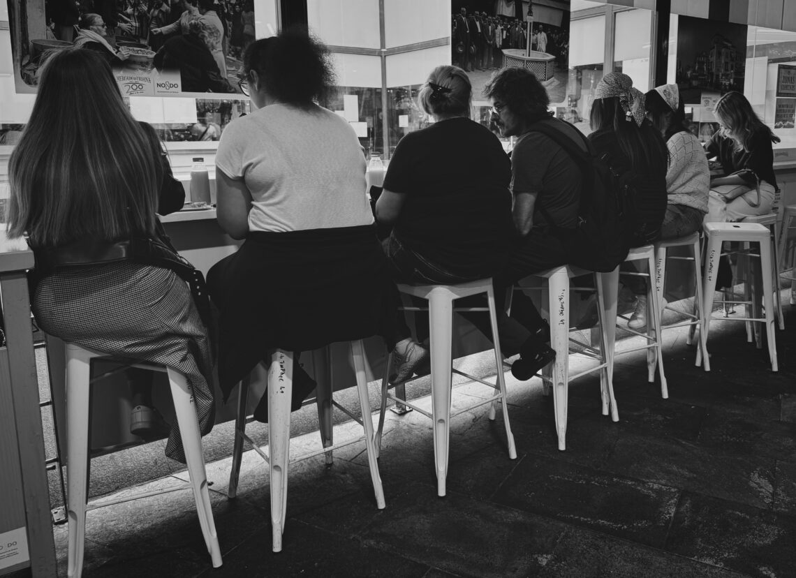 Mehrere Personen von hinten sitzend auf Barhockern an einer Theke im Mercado de Triana Sevilla, Schwarzweiß-Innenaufnahme