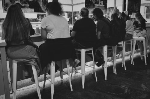 Mehrere Personen von hinten sitzend auf Barhockern an einer Theke im Mercado de Triana Sevilla, Schwarzweiß-Innenaufnahme