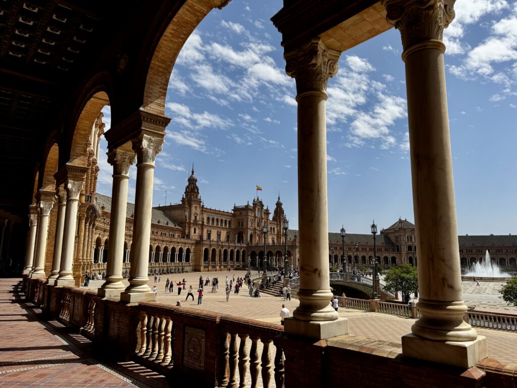 Blick durch den Arkadengang der Plaza de España in Sevilla auf den weitläufigen Platz mit Brunnen, Besuchern und dem Hauptgebäude unter blauem Himmel mit Wolken