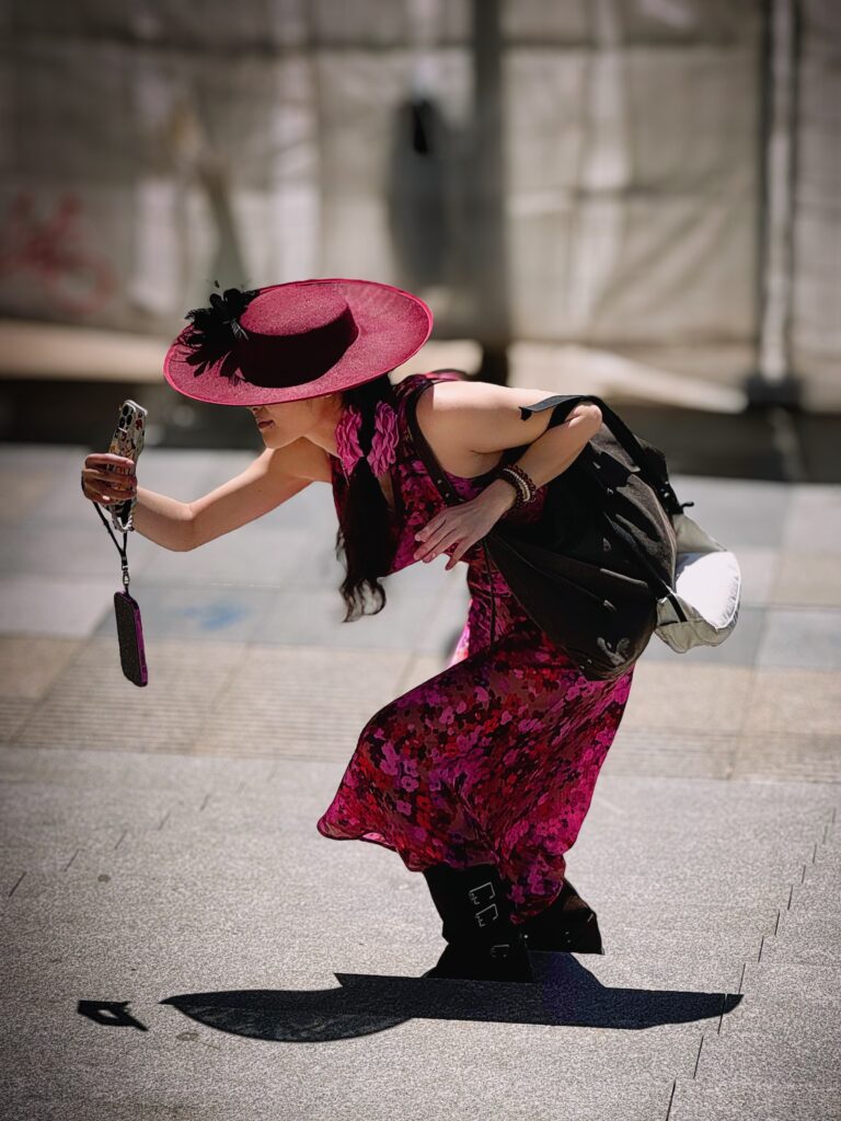 Frau in pinkem Kleid und großem Hut macht ein Selfie auf der Straße im Sonnenlicht – kreative Streetfotografie in kräftigen Farben