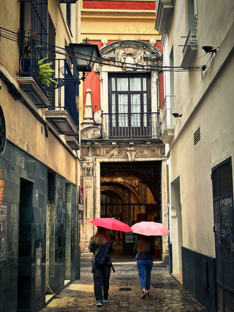 Zwei Personen mit roten Regenschirmen gehen durch eine enge Gasse mit historischer Fassade in Sevilla – Streetfotografie in Farbe mit architektonischem Fokus