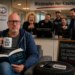 Genervter Mann sitzt beim Reifenwechsel im Toyota-Autohaus in Velbert im Wartebereich, während Mitarbeiter ihn lachend „Herr Steinfurth“ nennen.