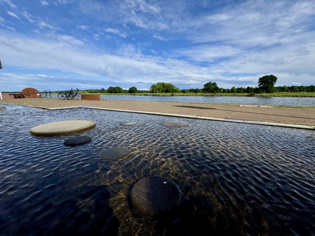 Ein flaches Wasserspiel mit runden Trittsteinen im Vordergrund an einer gepflasterten Promenade, dahinter der Fluss Maas und Fahrräder unter blauem Himmel.