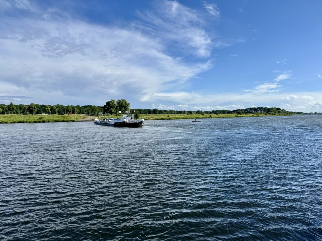 Eine Autofähre auf der Maas bei Kessel unter blauem Himmel mit weißen Wolken, im Hintergrund eine grüne Uferlandschaft mit Bäumen.