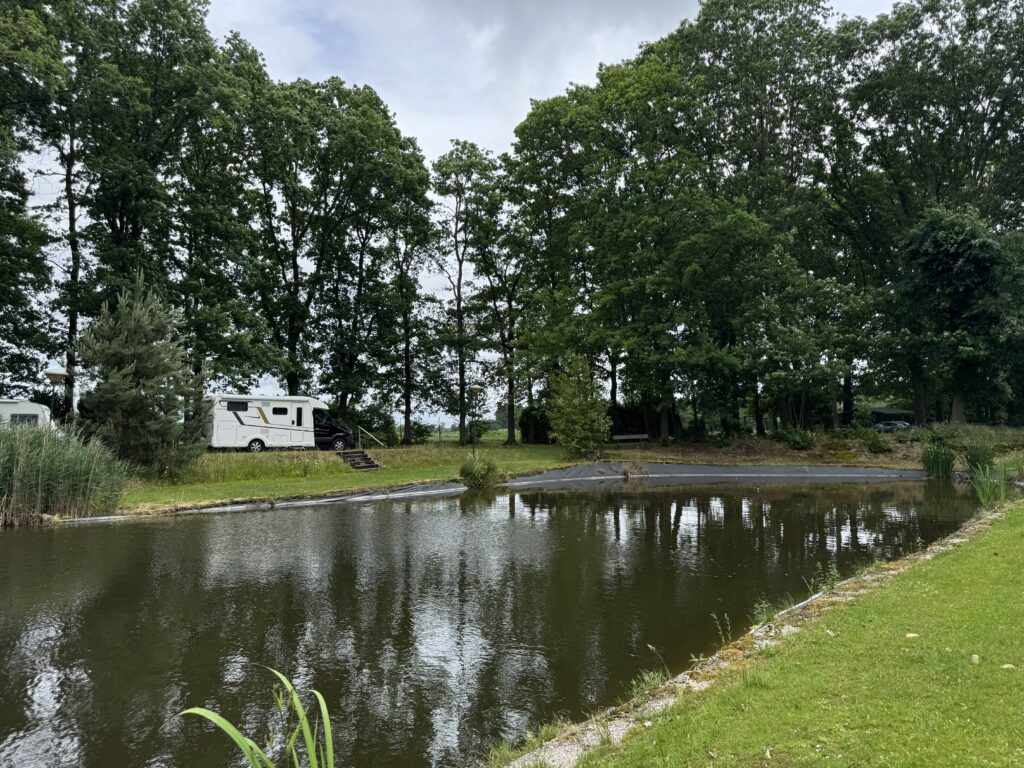 Ein weißes Wohnmobil parkt auf einer Wiese hinter einem Angelteich, umgeben von hohen, dichten Bäumen auf dem Campingplatz Boezeven.