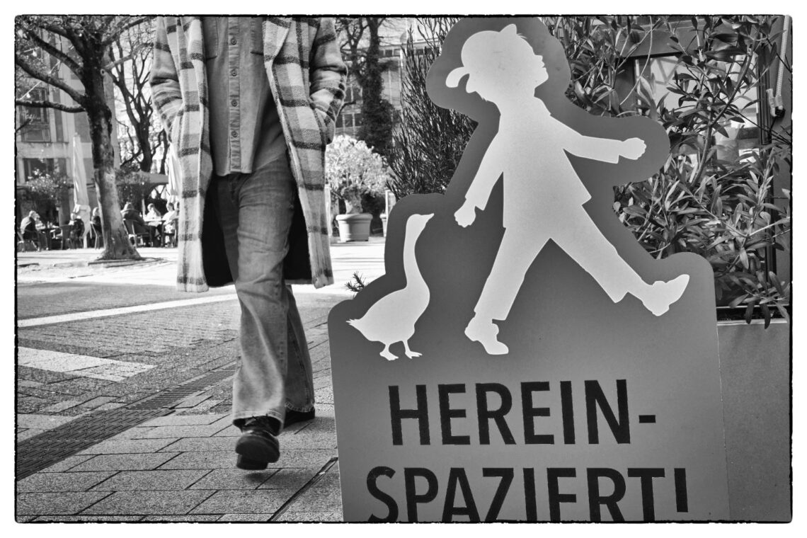 Streetfotografie in Wuppertal in Schwarzweiß: Ein roter Aufsteller mit einer Silhouette eines Kindes, einer Gans und der Aufschrift Hereinspaziert steht im Vordergrund. Dahinter läuft ein Passant in einem karierten Mantel vorbei, dessen Oberkörper abgeschnitten ist.
