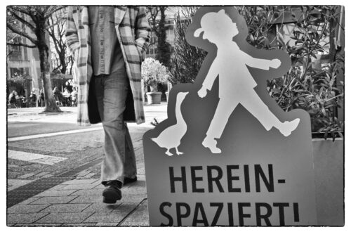Streetfotografie in Wuppertal in Schwarzweiß: Ein roter Aufsteller mit einer Silhouette eines Kindes, einer Gans und der Aufschrift Hereinspaziert steht im Vordergrund. Dahinter läuft ein Passant in einem karierten Mantel vorbei, dessen Oberkörper abgeschnitten ist.