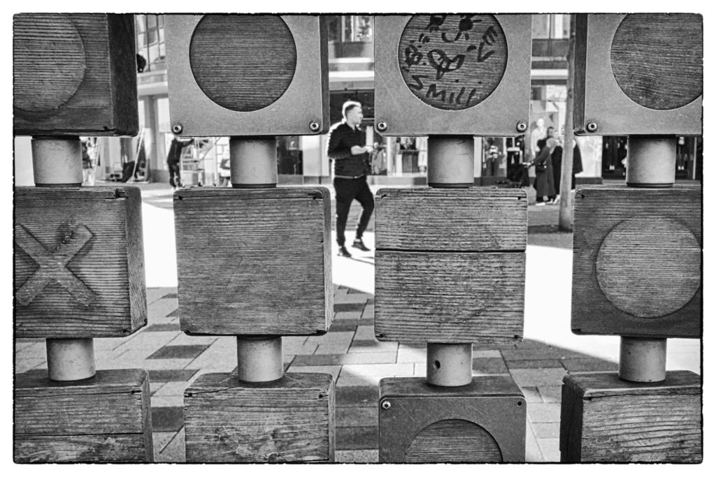 Eine hölzerne Tic-Tac-Toe-Spielstruktur auf einem öffentlichen Platz in Wuppertal. Durch die Lücken der Holzblöcke ist im unscharfen Hintergrund ein Passant beim Gehen in der Fußgängerzone zu sehen, aufgenommen in Schwarzweiß.