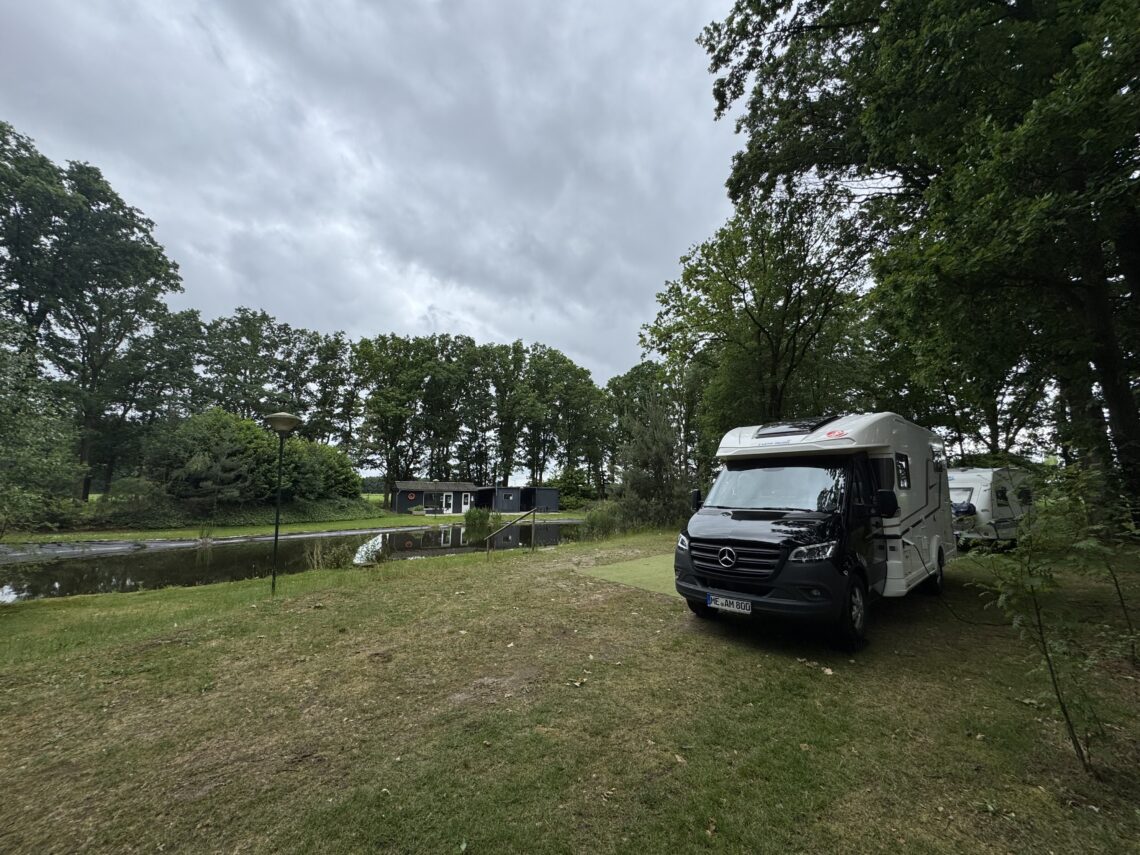 Ein weiß-schwarzes Wohnmobil steht auf einem Rasenstellplatz am Ufer eines Teichs auf dem Campingplatz in Kessel unter bewölktem Himmel.
