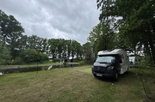Ein weiß-schwarzes Wohnmobil steht auf einem Rasenstellplatz am Ufer eines Teichs auf dem Campingplatz in Kessel unter bewölktem Himmel.