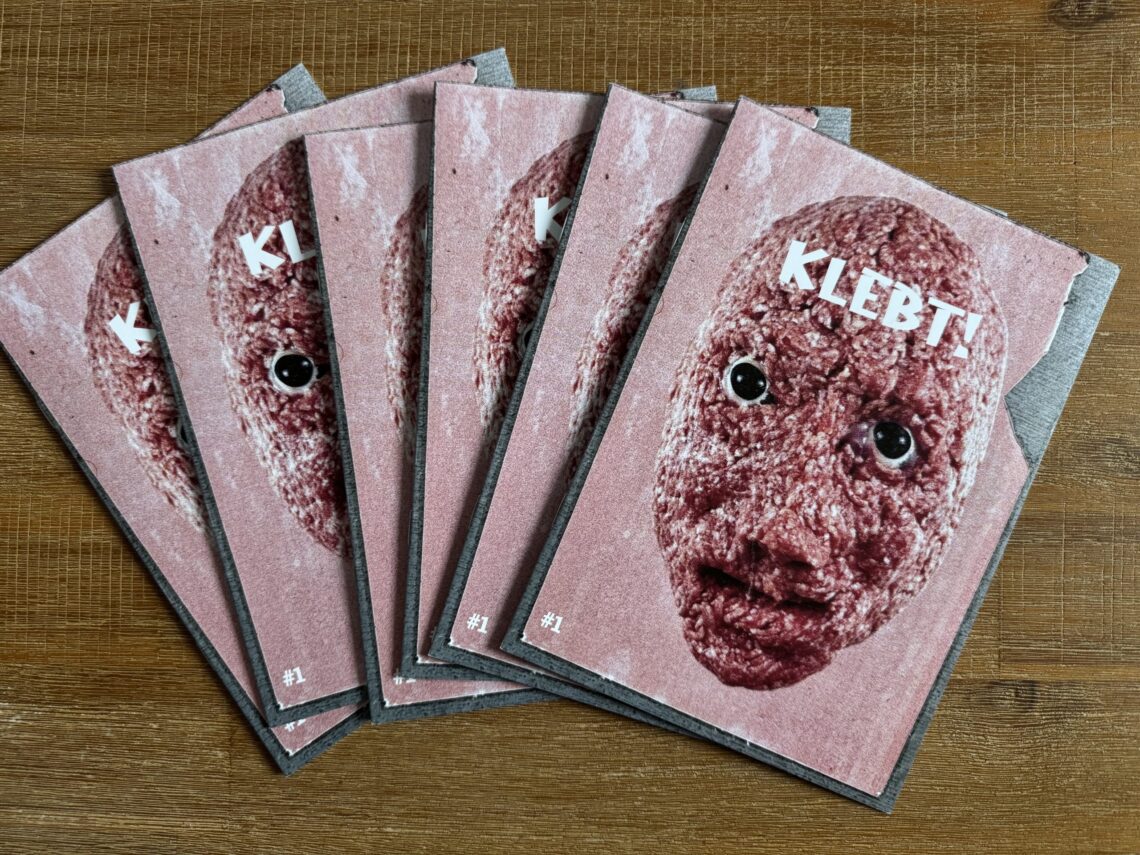 Stapel des gedruckten Fotozines KLEBT! Ausgabe 1 mit dem Titelmotiv eines Mettgesichts mit Kulleraugen auf rosafarbenem Hintergrund.