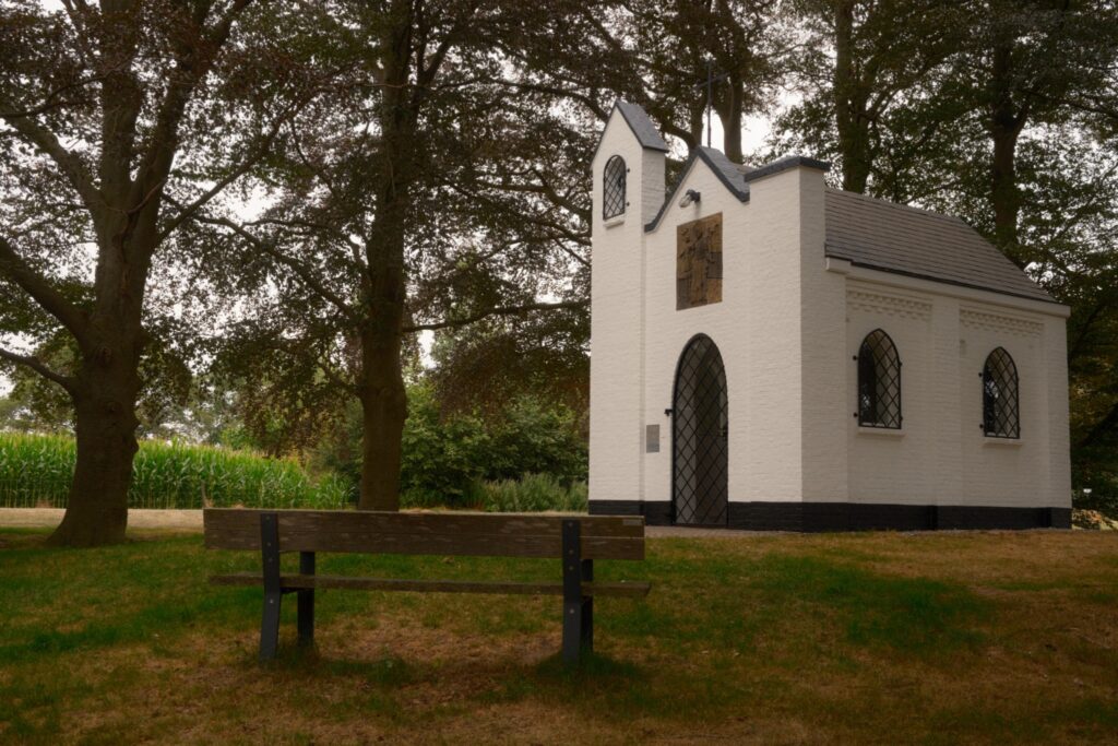 Kleine weiße Kapelle mit einer Holzbank im Schatten großer Bäume neben einem Maisfeld.