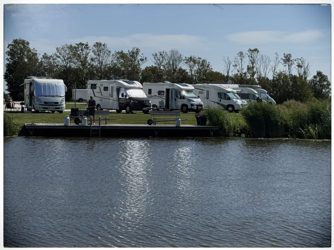 Aufnahme vom Wasser aus auf eine Reihe geparkter Wohnmobile am Ufer des Camperplaats aan de Ee bei Dokkum.