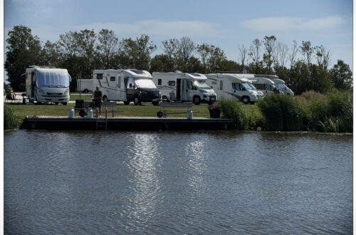 Aufnahme vom Wasser aus auf eine Reihe geparkter Wohnmobile am Ufer des Camperplaats aan de Ee bei Dokkum.