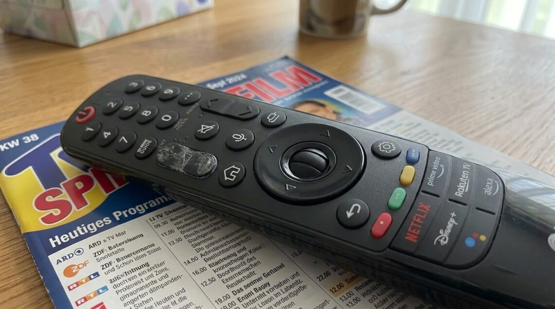 Nahaufnahme einer schwarzen LG Magic Remote TV-Fernbedienung, die auf einem geöffneten deutschen Fernsehprogrammheft ("TV Spielfilm", Ausgabe KW 38 / Sept 2024) auf einem Holztisch liegt. Die Fernbedienung zeigt Abnutzungsspuren am zentralen Scrollrad und verfügt über dedizierte Tasten für Streaming-Dienste wie Netflix, Disney+, Prime Video und Rakuten TV. Im unscharfen Hintergrund sind ein Taschentuchkarton und eine Tasse zu sehen.