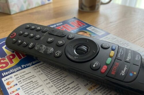 Nahaufnahme einer schwarzen LG Magic Remote TV-Fernbedienung, die auf einem geöffneten deutschen Fernsehprogrammheft ("TV Spielfilm", Ausgabe KW 38 / Sept 2024) auf einem Holztisch liegt. Die Fernbedienung zeigt Abnutzungsspuren am zentralen Scrollrad und verfügt über dedizierte Tasten für Streaming-Dienste wie Netflix, Disney+, Prime Video und Rakuten TV. Im unscharfen Hintergrund sind ein Taschentuchkarton und eine Tasse zu sehen.