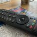 Nahaufnahme einer schwarzen LG Magic Remote TV-Fernbedienung, die auf einem geöffneten deutschen Fernsehprogrammheft ("TV Spielfilm", Ausgabe KW 38 / Sept 2024) auf einem Holztisch liegt. Die Fernbedienung zeigt Abnutzungsspuren am zentralen Scrollrad und verfügt über dedizierte Tasten für Streaming-Dienste wie Netflix, Disney+, Prime Video und Rakuten TV. Im unscharfen Hintergrund sind ein Taschentuchkarton und eine Tasse zu sehen.