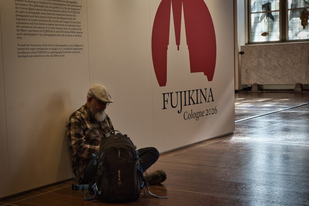 Ein Besucher mit Mütze und Rucksack sitzt entspannt am Boden neben der Aufschrift Fujikina Cologne 2026.