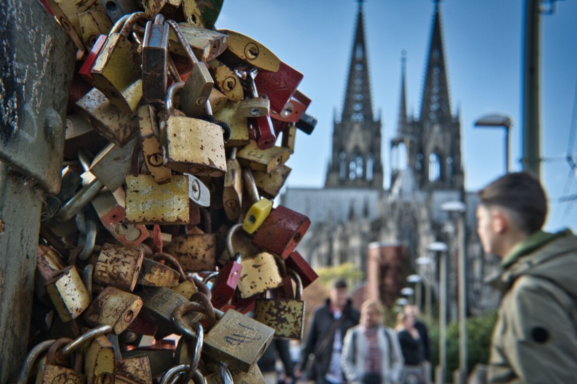 Nahaufnahme von Liebesschlössern an der Hohenzollernbrücke mit dem Kölner Dom im unscharfen Hintergrund.