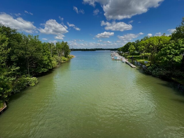 Blick auf das grüne Flussufer mit gepflegten Wiesen, Bäumen und einzelnen Bootsstegen an einem breiten Fluss. Im Vordergrund fährt ein Jet-Ski über das Wasser und hinterlässt eine weiße Spur. Der Himmel ist mit vielen Wolken bedeckt, dazwischen zeigen sich blaue Stellen. Im Hintergrund sind Felder und eine Bahntrasse zu sehen. Leukermeer, Radtour Kevelaer
