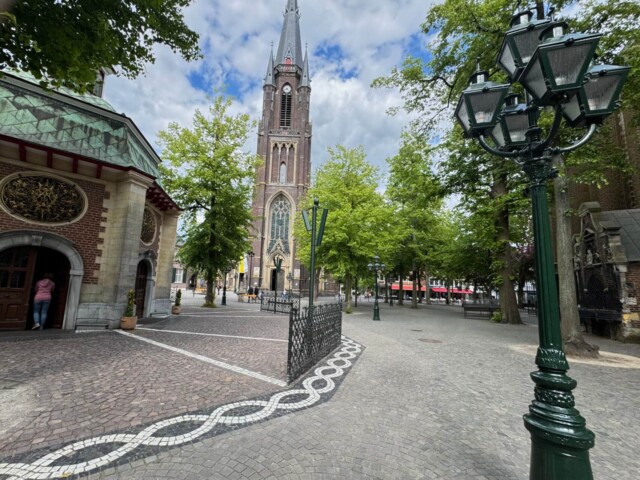 Kopfsteinplatz in Kevelaer mit Blick auf die neugotische Marienbasilika und die achteckige Gnadenkapelle im Vordergrund. Eine Frau betritt die Kapelle, daneben eine kunstvolle Laterne und gepflasterte Wege mit weißem Mosaikmuster. Der Platz ist von Bäumen gesäumt, im Hintergrund sind Straßencafés zu sehen, teils unter bewölktem Himmel. Radtour Keveler