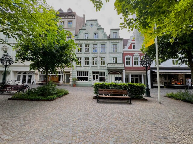 Historische Häuserfassaden in der Innenstadt von Kevelaer mit dem Gasthof „Zum weißen Kreuz“ im Zentrum. Vor dem Gebäude befindet sich ein kleiner, gepflasterter Platz mit Sitzbänken, Laternen und grüner Bepflanzung. Die Szene ist von Bäumen eingerahmt, im Hintergrund erkennt man weitere Geschäfte und Cafés. Radtour Kevelaer