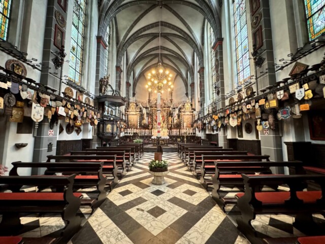 Blick ins Innere der Kerzenkapelle St. Michael in Kevelaer mit barock gestaltetem Altarraum, hohen Spitzbogenfenstern mit buntem Glas und dunklen Holzbänken. Die Seitenwände sind reich geschmückt mit Pilgerabzeichen, Wappen und Votivtafeln. Der Boden ist mit schwarz-weißem Marmor-Muster ausgelegt, eine Osterkerze steht im Mittelgang. Radtour Kevelaer