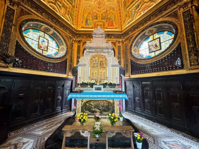 Blick auf den reich verzierten Altarraum in der Sakramentskapelle der Gnadenkapelle Kevelaer. Der Raum ist mit goldenen Wandmalereien, Mosaikfußboden und zwei großen ovalen Buntglasfenstern geschmückt. Vor dem Altar stehen Holzbänke und Blumenschmuck mit gelben und rosa Blumen, die eine Atmosphäre der Andacht und Ruhe erzeugen.