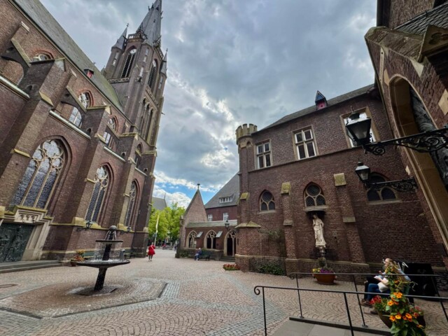 Innenhof der Marienbasilika Kevelaer mit Blick auf die neugotische Fassade und den hohen Kirchturm. Umgeben von Backsteingebäuden mit Spitzbogenfenstern und kleinen Türmen. In der Mitte des gepflasterten Platzes steht ein Brunnen, rechts eine Marienstatue in einer Nische. Blumen schmücken den Vorplatz, eine Frau sitzt auf einer Bank. Der Himmel ist wolkenverhangen.