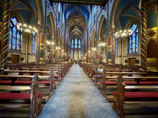 Innenansicht der farbenprächtigen Marienbasilika in Kevelaer mit reich verzierten Säulen, blauen Decken mit goldenen Sternmustern und farbigen Glasfenstern. Der Blick führt durch das Hauptschiff zum goldenen Hochaltar. Auf beiden Seiten stehen kunstvoll geschnitzte Holzbänke mit roten Polstern. Eine Frau sitzt betend in einer der vorderen Reihen.