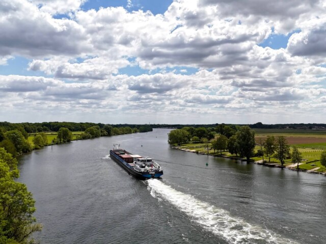 Ein Frachtschiff fährt auf einem breiten Fluss durch eine grüne Landschaft mit Bäumen und Feldern unter einem teils bewölkten Himmel. Am Ufer sind Spazierwege, Liegewiesen und ein kleiner Hafenbereich zu sehen. Das Schiff hinterlässt eine weiße Heckwelle im Wasser. Radtour Kevelaer