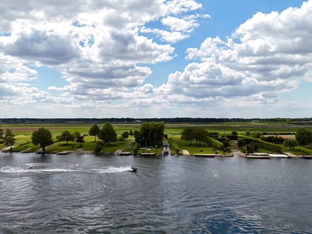 Blick auf das grüne Flussufer mit gepflegten Wiesen, Bäumen und einzelnen Bootsstegen an einem breiten Fluss. Im Vordergrund fährt ein Jet-Ski über das Wasser und hinterlässt eine weiße Spur. Der Himmel ist mit vielen Wolken bedeckt, dazwischen zeigen sich blaue Stellen. Im Hintergrund sind Felder und eine Bahntrasse zu sehen. Radtour Kevelaer