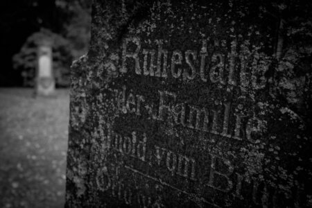 Nahaufnahme eines stark verwitterten Grabsteins mit der erkennbaren Inschrift „Ruhestätte der Familie Arnold vom Broch“. Der Stein ist mit Flechten und Moos überzogen. Im Hintergrund steht ein weiterer Grabstein, unscharf und etwas entfernt, eingebettet in herbstliche Natur. Stiller Park Velbert