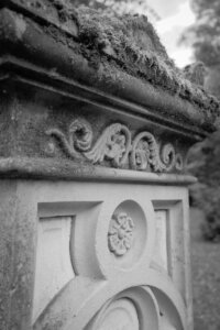 Detailansicht der ornamentalen oberen Kante eines historischen Grabmals. Gut erhaltene florale Steinverzierungen und kreisförmige Blumenmotive sind erkennbar, teilweise mit Moos überwuchert. Das Foto zeigt die handwerkliche Steinmetzkunst vergangener Jahrhunderte. Seiler Park Velbert