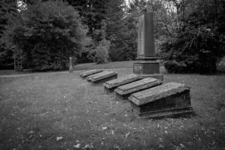 Gruppe von fünf schräg liegenden, flachen Grabsteinen aus dunklem Stein, in einer Reihe angeordnet. Dahinter ein großes, aufrecht stehendes Denkmal. Die Szenerie ist von dichter Vegetation umgeben, das Gras wirkt leicht verwildert, herbstliche Blätter bedecken den Boden. Stiller Park Velbert.