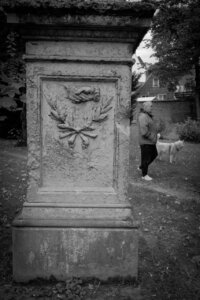 Ein reliefverzierter Grabstein mit Händedruck-Motiv und Lorbeerzweigen im Vordergrund. Im Hintergrund spaziert ein älterer Mann mit hellem Hund auf einem Weg vorbei. Der Kontrast zwischen bewegtem Alltag und stiller Ewigkeit ist spürbar. Stiller Park Velbert.