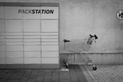 Verlassener Einkaufswagen vor Packstation an Hauswand, minimalistische Streetfotografie Schwarzweiß, Alltagsszene in deutscher Kleinstadt