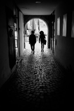 Zwei Personen als Silhouetten in dunklem Durchgang mit hellem Torbogen, Schwarzweiß-Streetfotografie, starker Hell-Dunkel-Kontrast, Kopfsteinpflasterboden