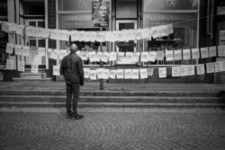 Person von hinten betrachtet Schaufenster eines Goldschmieds mit zahlreichen aufgehängten Zetteln, dokumentarische Schwarzweiß-Streetfotografie, Kopfsteinpflaster