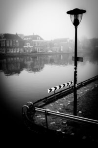 Laterne und Verkehrsschilder am Flussufer, nebelverhüllte Häuser spiegeln sich im Wasser in Sneek.